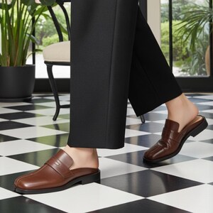 May include: Brown leather slip-on loafers with a low heel. The shoes have a classic design with a strap across the top. The person is wearing black pants. The floor has a black and white checkered pattern.