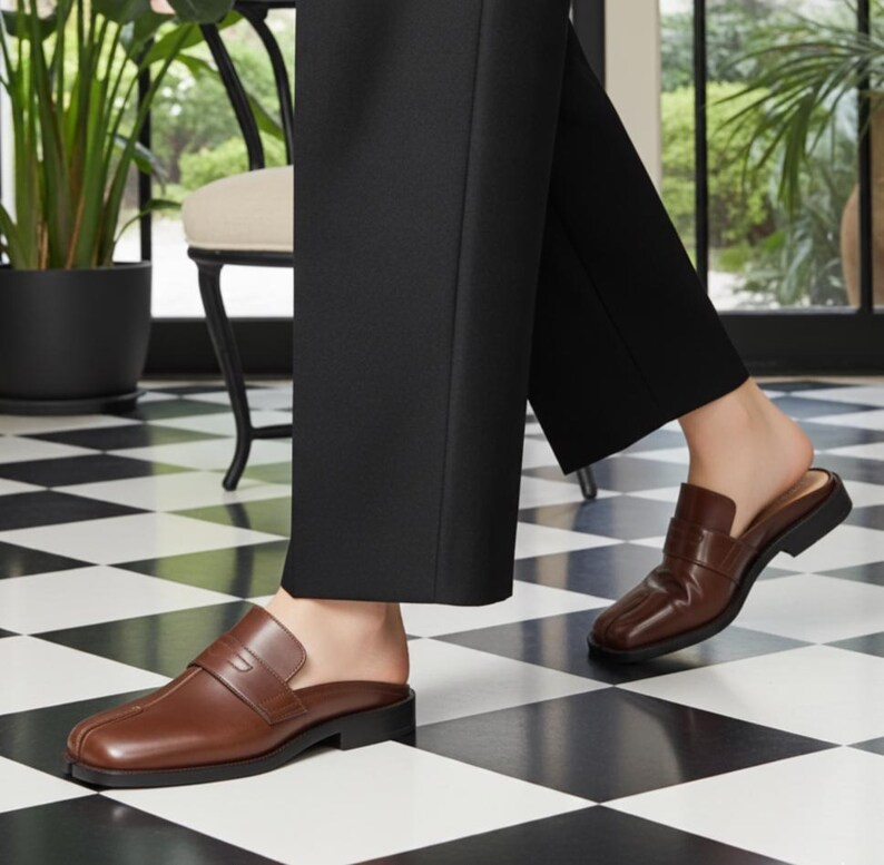 May include: Brown leather slip-on loafers with a low heel. The shoes have a classic design with a strap across the top. The person is wearing black pants. The floor has a black and white checkered pattern.
