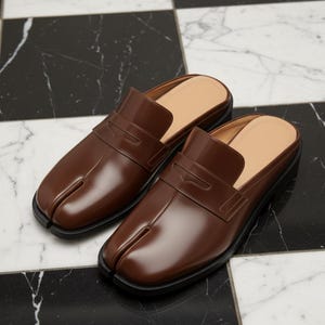 May include: A pair of brown leather slip-on shoes with a split-toe design. The shoes have a square toe, a strap across the top, and a black sole. The shoes are displayed on a black and white checkered surface.
