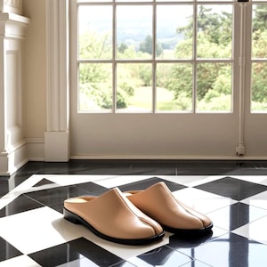 May include: A pair of beige leather slip-on shoes with a split toe design and black soles. The shoes are set on a black and white checkered floor, with a window and outdoor scenery in the background.