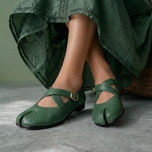 May include: A pair of dark green leather split-toe shoes featuring a low heel, a rounded toe box with a distinct vertical slit, and crisscrossing straps across the top of the foot secured with a small, circular brass-toned metal buckle.