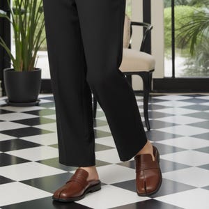 May include: Brown leather split-toe slip-on shoes with a low heel. The shoes have a strap across the top and are worn with black pants. The floor is a black and white checkered pattern.