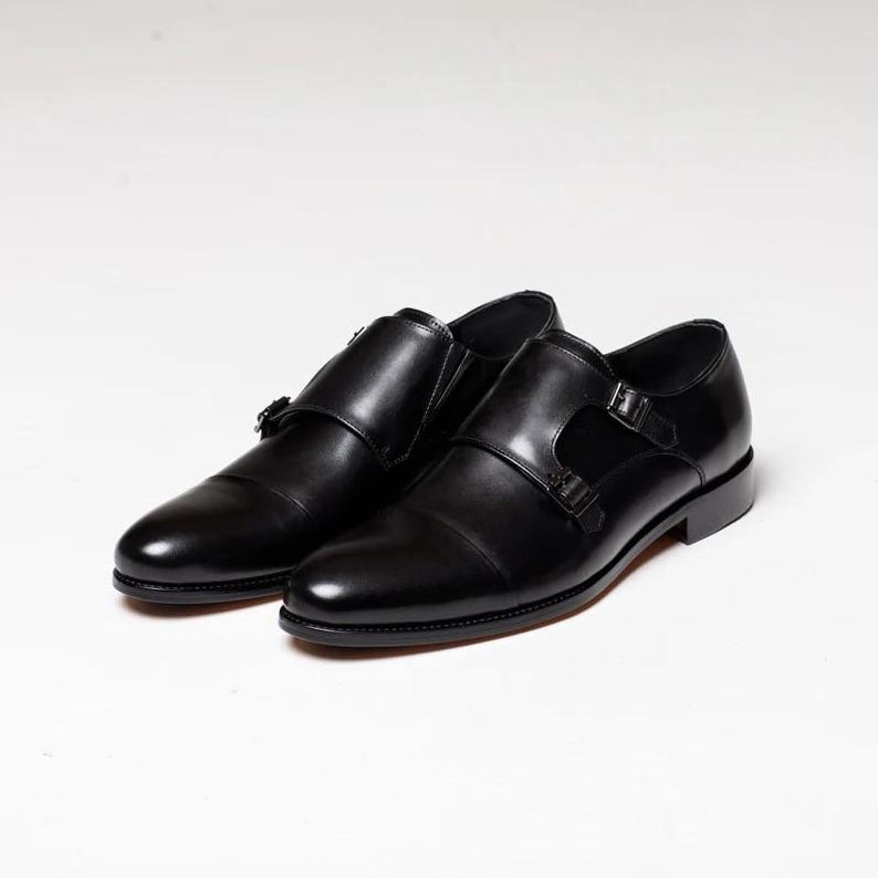 Puede incluir: Un par de zapatos de vestir de cuero negro con doble hebilla monje. Los zapatos tienen un dise&ntilde;o cl&aacute;sico con punta puntiaguda y cierre de hebilla.