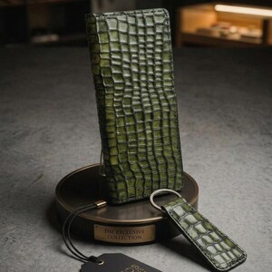 Pode incluir: Conjunto de carteira e porta-chaves em couro verde com textura de crocodilo. A carteira está em pé sobre um suporte redondo com uma placa dourada que diz "THE EXCLUSIVE COLLECTION". Uma etiqueta preta com texto branco está pendurada abaixo.