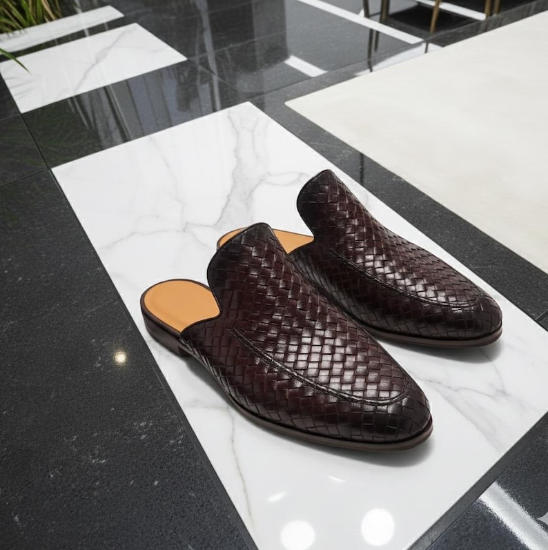 May include: A pair of dark brown woven leather slip-on shoes. The shoes have a rounded toe and a low heel. The interior is a light tan color. The shoes are displayed on a white marble surface.