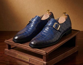May include: A pair of blue leather monk strap shoes with a crocodile skin texture and a gradient finish, featuring a gold-toned metal buckle and a wooden shoe tree inside each shoe, displayed on a tiered wooden stand with a Master Craftsman plaque.