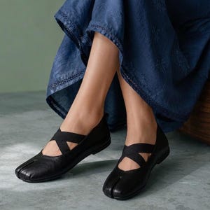 May include: A pair of black leather split-toe flats featuring a unique tabi-style design with a distinct separation between the big toe and the other toes. The shoes include black elastic crisscross straps over the instep and a flat sole.