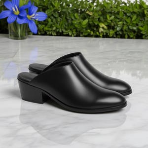 Può includere: Un paio di mules in pelle nera con tacco basso a blocco. Le scarpe hanno la punta arrotondata e un design slip-on. I tacchi sono alti circa 5 cm.