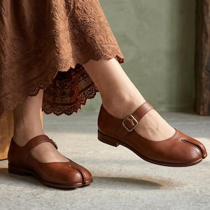 May include: A pair of brown leather Mary Jane shoes featuring a distinctive split-toe design, a low heel, and a thin adjustable strap across the instep secured with a small brass-toned metal buckle. The shoes have a smooth finish and rounded toe.