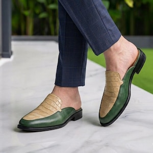 May include: A pair of backless loafers featuring a two-tone design with a beige crocodile-embossed leather vamp and a smooth dark green leather body, paired with navy blue plaid trousers. The shoes have a low heel and a pointed toe silhouette.
