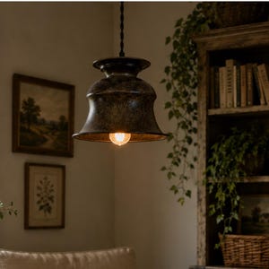 May include: A dark metal bell-shaped pendant light fixture with a warm glowing bulb suspended from a black cord, featuring a wide rim and a smooth, weathered finish that hangs above a beige upholstered armchair with a floral patterned pillow.