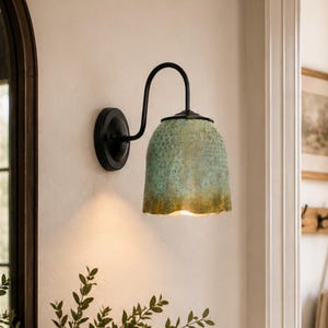May include: A wall-mounted light fixture featuring a curved black metal arm and a bell-shaped glass shade with a textured, mottled green and gold finish. The lamp emits a warm glow from beneath the shade, which has a scalloped bottom edge.