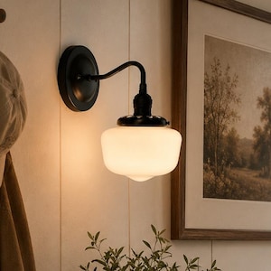 May include: A wall-mounted light fixture featuring a matte black metal base and a curved arm supporting a white, bell-shaped glass shade. The lamp emits a warm, soft glow, illuminating the surrounding area with a gentle, diffused light.