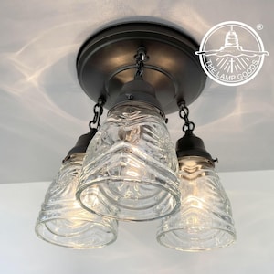 May include: A three-light ceiling fixture with a black metal canopy and three clear glass shades with a textured pattern. The fixture is hanging from the ceiling and the lights are on.