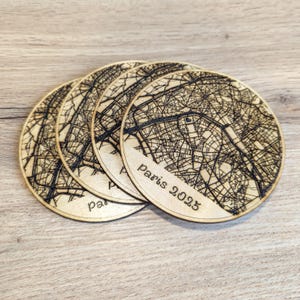 May include: Four round wooden coasters with a map of Paris etched into them. The coasters are light brown with a dark brown map design. The text "Paris 2023" is visible on one of the coasters.