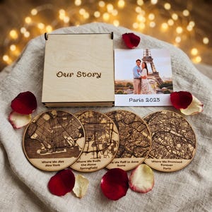 May include: A light-colored wooden box with the text Our Story, a square photograph of a couple in front of the Eiffel Tower with the text Paris 2023, and four circular wooden coasters featuring engraved city maps and text labels for New York, Springfield, Rome, and Paris.