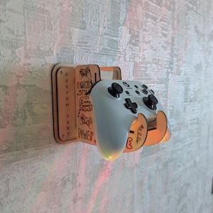 May include: A wooden wall-mounted gaming controller holder with a light gray controller. The holder has etched text and graphics, including "CUSTOM TEXT" and "POWER". The controller is held in place by two curved supports.
