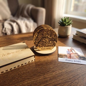 Peut inclure: Un ensemble de dessous de verre circulaires en bois avec une carte de Paris gravée au laser en noir et le texte Paris 2025, présentés sur un support en bois. Une boîte en bois avec le texte Where we met et une photo carrée sont à côté.
