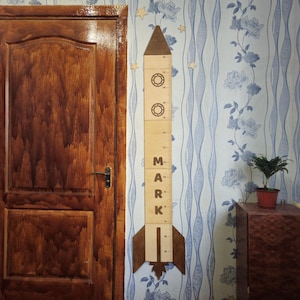May include: A wooden rocket-shaped height chart with the name "MARK" printed on it. The chart is light brown with darker brown accents and measurement markings. It is mounted on a wall with blue floral wallpaper.