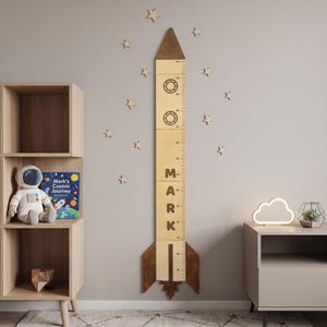 May include: A wooden rocket ship growth chart featuring a dark brown pointed top and base, a light wood body with two circular porthole cutouts, and a vertical measurement scale. The name MARK is printed in large dark letters along the center.