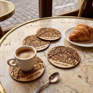 May include: Four round wooden coasters featuring a dark engraved map of Paris and the text Paris 2025. One coaster supports a white ceramic espresso cup filled with coffee, while a croissant sits on a small white plate nearby.