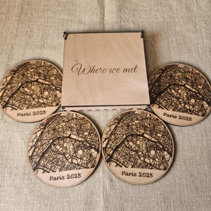 May include: A light-colored wooden square box with the engraved text Where we met, accompanied by four circular wooden coasters. Each coaster features a laser-engraved map of Paris with the text Paris 2025 at the bottom edge.