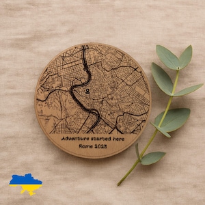 Può includere: Un sottobicchiere rotondo in legno con una mappa di Roma e la scritta "Adventure started here Rome 2023". Il sottobicchiere è marrone chiaro con dettagli della mappa neri. Un rametto di foglie verdi è accanto al sottobicchiere.