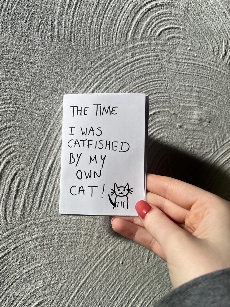 May include: A white sticky note with black handwritten text that reads "THE TIME I WAS CATFISHED BY MY OWN CAT!" A small cat drawing is next to the text. The note is held by a hand with red nail polish.