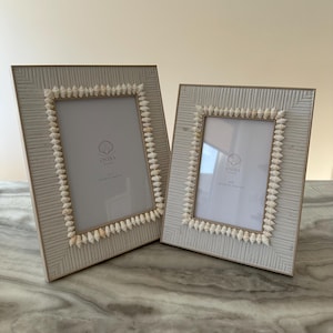 Könnte beinhalten: Zwei weiße Bilderrahmen mit einem dekorativen Muschelrand. Die Rahmen sind aus Holz und haben eine hellbraune Oberfläche. Die Rahmen sind beide leer und bereit, mit Fotos gefüllt zu werden.