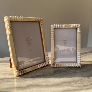 Könnte beinhalten: Zwei weiße Bilderrahmen mit Muschel-Design. Die Rahmen sind aus Holz und haben eine natürliche Oberfläche. Der größere Rahmen hat den Text "Ostra Studio" auf der Innenseite gedruckt. Der kleinere Rahmen hat den Text "Ostra Studio" auf der Innenseite gedruckt.