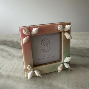 May include: A small, ombre picture frame with a seashell design. The frame is pink, green, and white and has a seashell design around the edges. The frame is 2.5 x 2.5 inches and is approximately 6 cm. The frame is made by OSTRa STUDIO.
