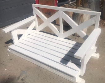 May include: White wooden swing with a criss-cross design on the back. The swing has a slatted seat and is made of unfinished wood.