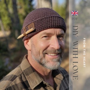 May include: A dark brown crocheted beanie with a folded brim, featuring a small brown leather patch on the side that holds a light-colored wooden pencil. The text on the right side reads PDF DIY WITH LOVE FROM THE HEART.