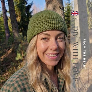 Peut inclure: Un bonnet crocheté vert olive avec un bord replié. Le texte sur le côté droit indique : PDF DIY WITH LOVE FROM THE HEART, accompagné d'une petite icône de drapeau du Royaume-Uni. Le bonnet présente un motif de mailles dense et uniforme.