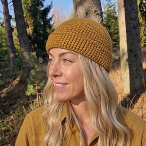 Peut inclure: Un bonnet jaune moutarde avec un large revers replié. Le bonnet est réalisé avec un point de crochet dense et uniforme, créant une surface texturée. Il présente une calotte arrondie et une coupe ajustée autour de la tête.
