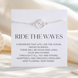 Pode incluir: Uma pulseira prateada com um pingente de onda, exibida em um cartão branco. O cartão mostra o texto "RIDE THE WAVES" e um texto inspirador. O fundo inclui uma concha e tecido.