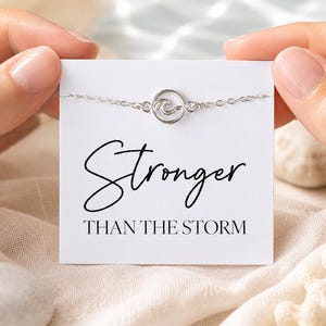Peut inclure: Un bracelet argenté avec un motif de vague est présenté sur une carte blanche. La carte porte l'inscription "Stronger Than The Storm" en écriture noire. Le bracelet est tenu par deux mains.