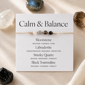 Puede incluir: Una pulsera de cadena dorada con cuatro cuentas de piedras preciosas: blanca, gris, marrón y negra. La pulsera se muestra en una tarjeta blanca con el texto "Calm & Balance" y los nombres de las piedras. Hay piedras adicionales alrededor de la tarjeta.