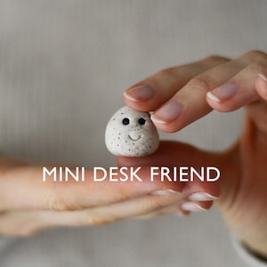 Op de afbeelding: Een kleine, witte, eivormige figuur met zwarte spikkels en een lachend gezicht. De tekst "MINI DESK FRIEND" staat onder de figuur. De figuur wordt tussen twee vingers gehouden.