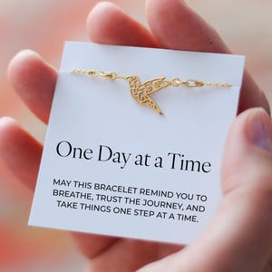 May include: A gold-colored hummingbird bracelet is displayed on a white card. The card reads "One Day at a Time" with an inspirational message. The bracelet is held in a person's hand.