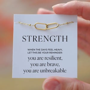 Op de afbeelding: Een goudkleurige kettingarmband met twee ovale, aan elkaar gekoppelde bedels. De armband wordt getoond op een witte kaart met het woord "STRENGTH" en de inspirerende tekst: "you are resilient, you are brave, you are unbreakable."