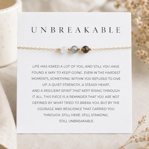 May include: A delicate gold chain bracelet featuring three small, faceted gemstone beads in white, grey, and dark brown, centered on a white display card. The card displays the text UNBREAKABLE and a paragraph of inspirational English text.