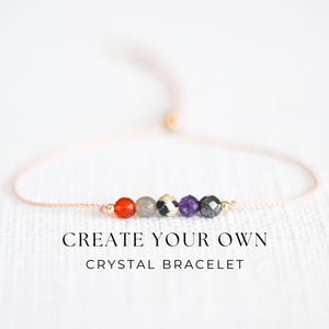 May include: A delicate crystal bracelet featuring a series of colorful gemstone beads, including red, gray, black and purple, strung on a light pink cord. The text "CREATE YOUR OWN CRYSTAL BRACELET" is displayed below the bracelet.