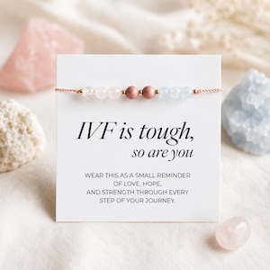 May include: A beaded bracelet on a white display card featuring the text "IVF is tough, so are you" and "WEAR THIS AS A SMALL REMINDER OF LOVE, HOPE, AND STRENGTH THROUGH EVERY STEP OF YOUR JOURNEY." The bracelet has faceted clear, pink, and blue beads.