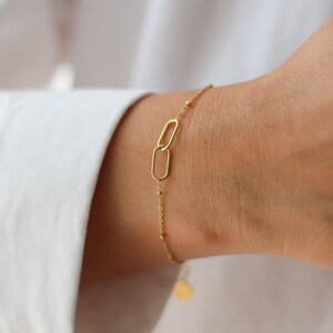 May include: A delicate gold-colored bracelet featuring two linked oval charms as a focal point. The bracelet has a fine chain with small, evenly spaced beads. A small, round charm hangs from the end of the chain.