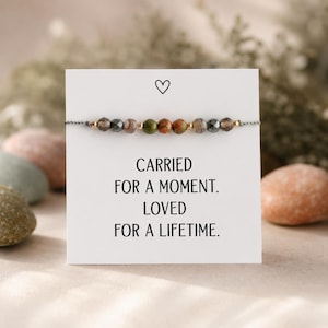 Pode incluir: Uma pulseira de contas com pedras em tons terrosos, enfiadas num cordão cinzento. Apresentada num cartão branco com um coração e o texto "CARRIED FOR A MOMENT. LOVED FOR A LIFETIME."
