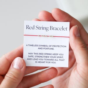Pode incluir: Uma pulseira de fio vermelho em um cartão branco. O cartão diz "Red String Bracelet" e inclui texto sobre proteção e fortuna. A pulseira apresenta pequenas contas douradas.