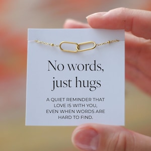 Puede incluir: Un brazalete dorado con dos dijes ovalados unidos se muestra en una tarjeta blanca. La tarjeta tiene el texto "No words, just hugs" y un mensaje sobre el amor. El brazalete es sostenido por una mano.