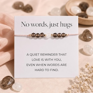 May include: A bracelet featuring brown and white beads strung on a pink cord. The bracelet is presented on a white card with the text "No words, just hugs" and a message about love. The background includes crystals and flowers.