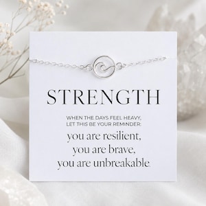 Pode incluir: Uma pulseira prateada com um berloque de onda é exibida em um cartão branco. O cartão diz "STRENGTH" e inclui o texto "you are resilient, you are brave, you are unbreakable."
